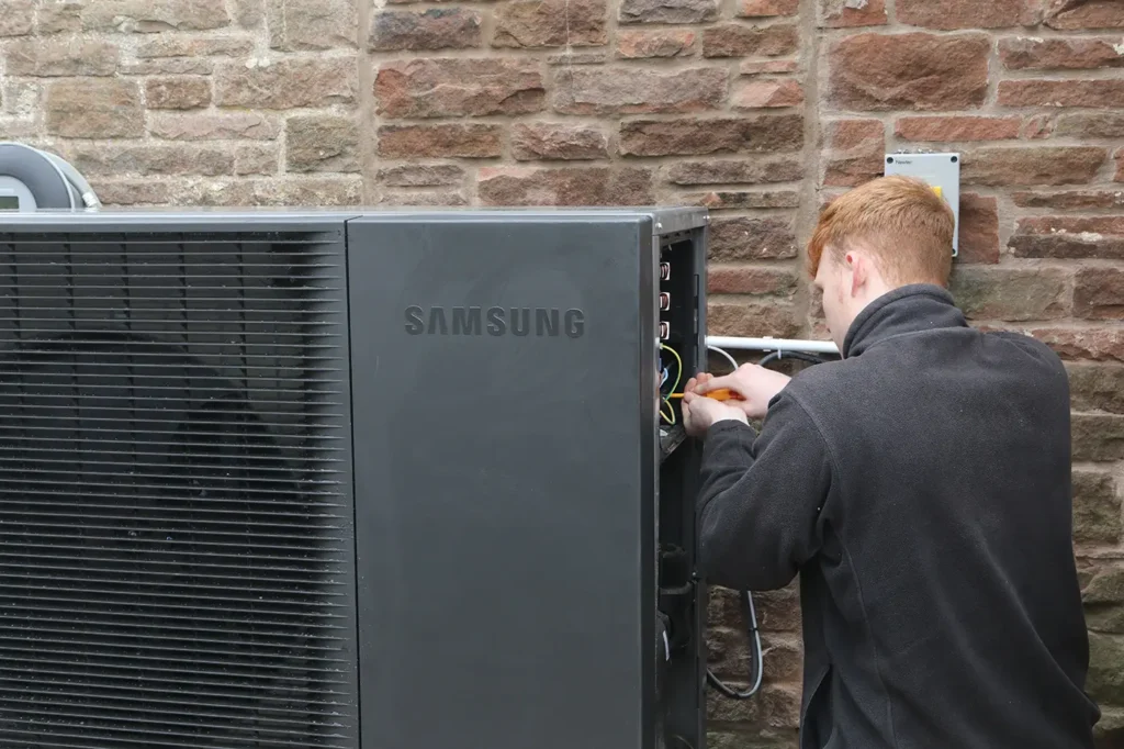 Technician servicing a Samsung air source heat pump installed against a stone wall, ensuring efficient operation and performance.