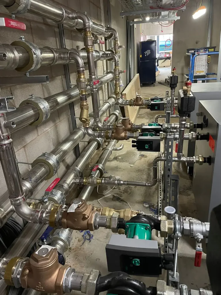 Close-up view of the piping and mechanical components behind a commercial ground source heat pump system. The image shows stainless steel pipes, valves, circulator pumps, and control units, forming part of the heat distribution and regulation system in a mechanical room.