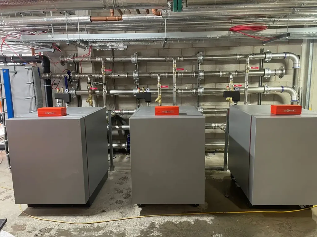 Three large commercial ground source heat pumps installed in a mechanical room, with extensive stainless steel piping, valves, and gauges mounted on the wall behind them.