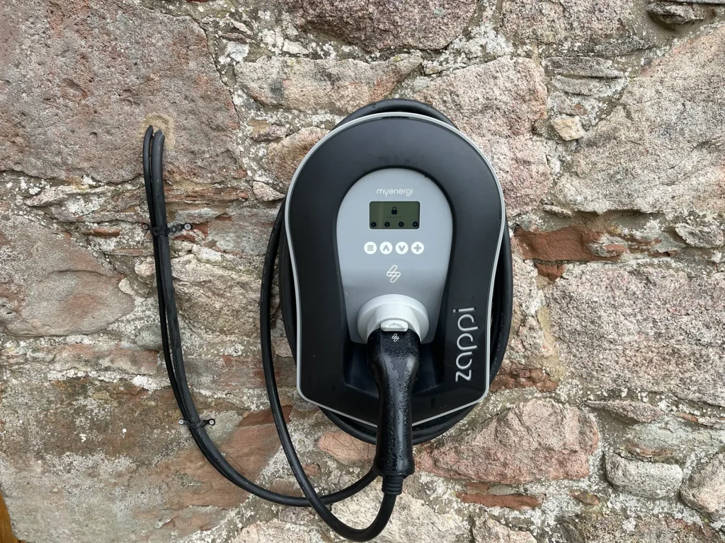 Front view of a Zappi electric car charger mounted on a stone wall, featuring a coiled charging cable and digital display for efficient EV charging.