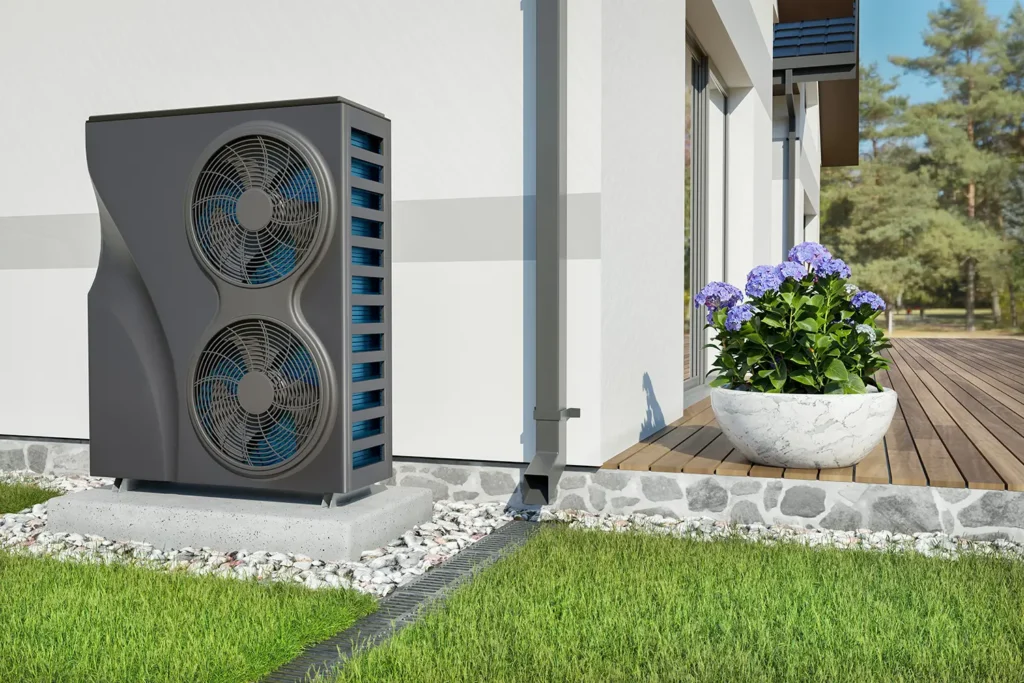 Modern air source heat pump installed outside a contemporary home, placed on a concrete base with surrounding landscaping, including a planter with flowers and a wooden deck.