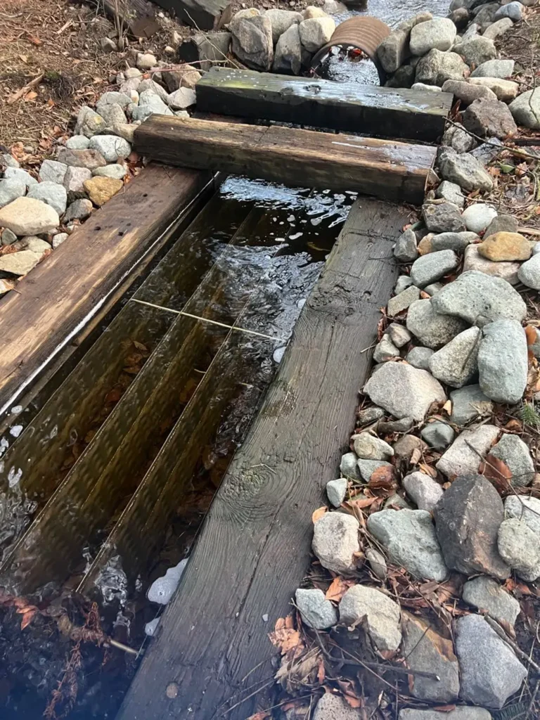 A water intake system for a water source heat pump, featuring a structured wooden channel directing water flow. The setup includes wooden beams and a rock bed to stabilize the intake and filter debris. Water flows from a culvert pipe into the collection area, ensuring a steady source for heat exchange.