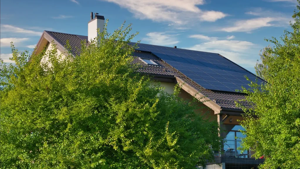 A modern house with solar panels installed on its sloped roof, surrounded by lush green trees under a blue sky with scattered clouds.